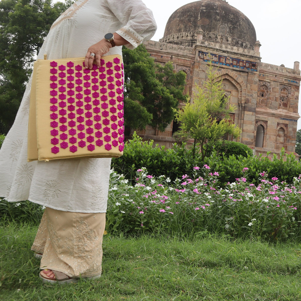 Sonja carrying the rolled LotuSun mat past a flower bed, a Delhi tomb in the background